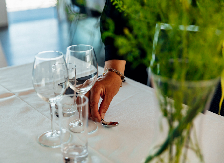 En person dukar ett bord med vinglas och bestick i en ljus konferenslokal på Västerviks Stadshotell, med gröna växter som dekoration.