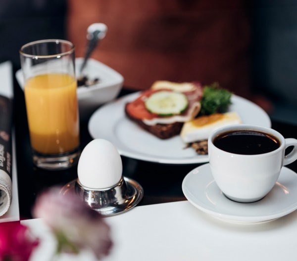 En närbild av en hotellfrukost på Västerviks Stadshotell, med en kopp kaffe, ett glas apelsinjuice, en tallrik med smörgås och ett kokt ägg.